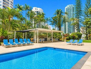 Ein Swimmingpool mit Liegestühlen, einem überdachten Sitzbereich und hohen Gebäuden im Hintergrund.