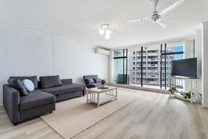 Ein modernes Wohnzimmer mit einem grauen Ecksofa, einem weißen Couchtisch, einem Deckenventilator und einem großen Fenster mit Blick auf ein Gebäude.