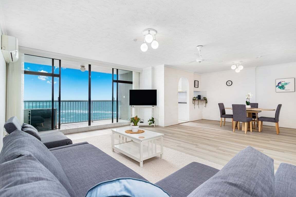 A modern living room with a large TV, a dining area, and a balcony with a view of the ocean.