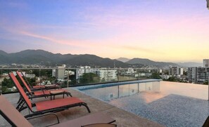 Piscina infinita no terraço com espreguiçadeiras, com vista para a paisagem urbana e montanhas.