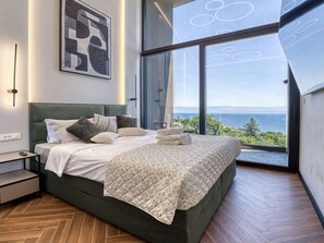 Une chambre moderne avec un grand lit, une table de chevet, une lampe fixée au mur et une grande fenêtre donnant sur la mer.