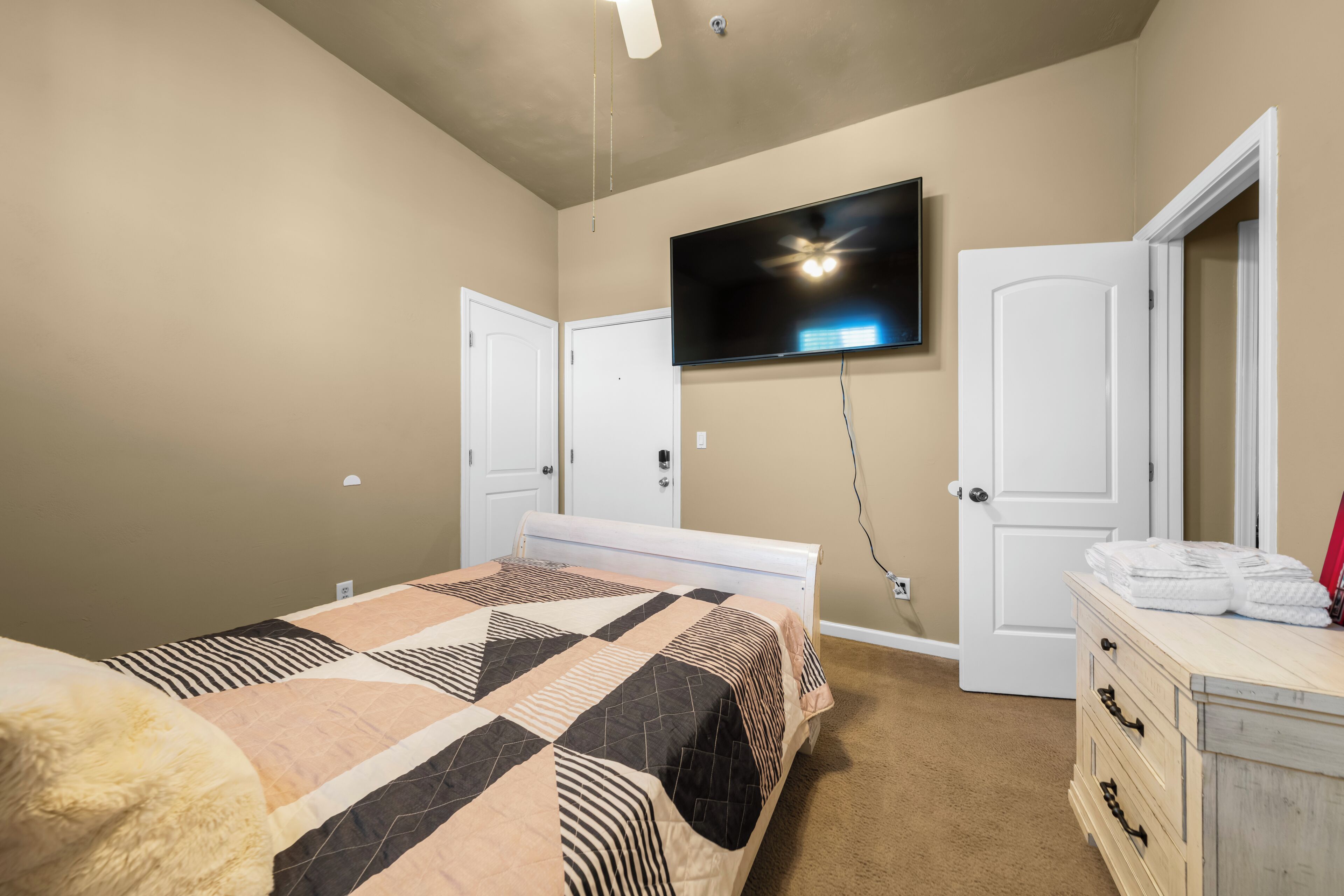 A bedroom with a bed, a TV mounted on the wall, a dresser, and two doors.