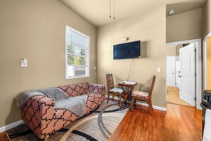 A living room with a sofa, a TV mounted on the wall, a dining table with chairs, and a door leading to another room.