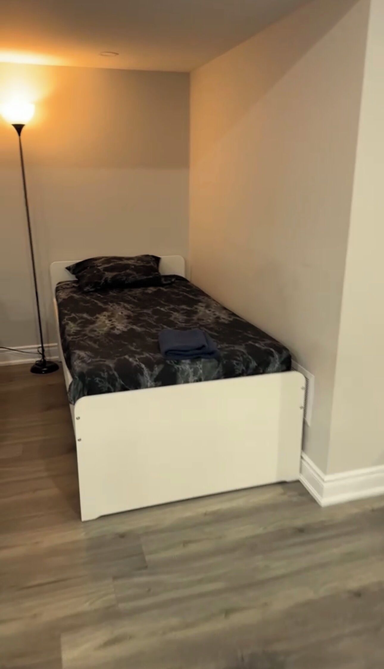 A single bed with a white frame and a dark patterned bedspread, a floor lamp, and a light-colored wall.