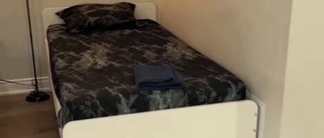 A single bed with a white frame and a dark patterned bedspread, a floor lamp, and a light-colored wall.