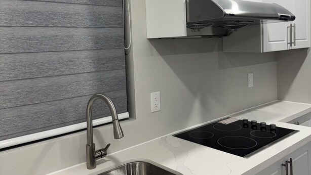 A modern kitchen with a stainless steel sink, a black stovetop, white cabinets, and a window with a grey blind.