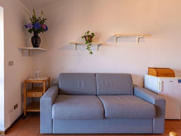 A living room with a grey sofa, a small wooden table with a lantern, a white refrigerator, and two wall-mounted shelves with plants. - Apartment Niki by Interhome (Domaso)