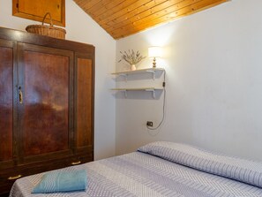 Een slaapkamer met een houten garderobe, een bed met beddengoed met een patroon, een wandplank met een lamp, en een mand aan de muur.