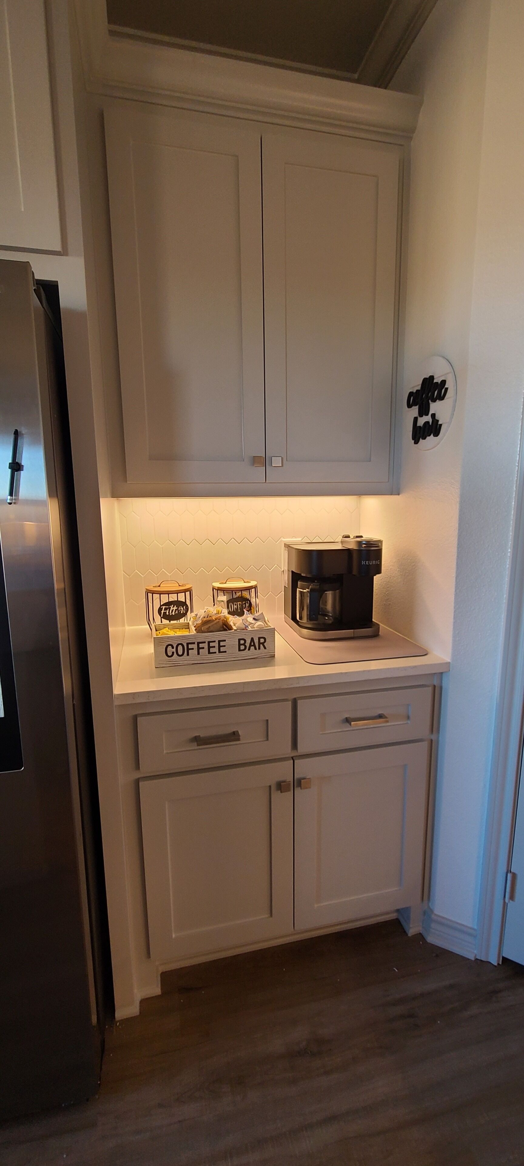 A kitchen with white cabinets, a coffee bar, a coffee maker, and a refrigerator.