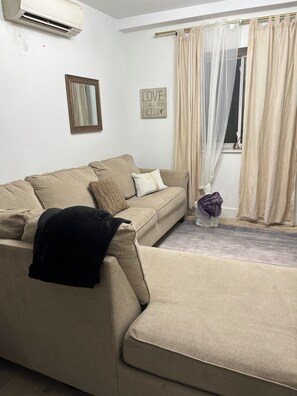 A living room with a beige sofa, a mirror, a 'LOVE IS ALL YOU NEED' sign, and beige curtains. - Enjoyable 1-bedroom apartment with AC 
WiFi in charming Corona. (Corona)