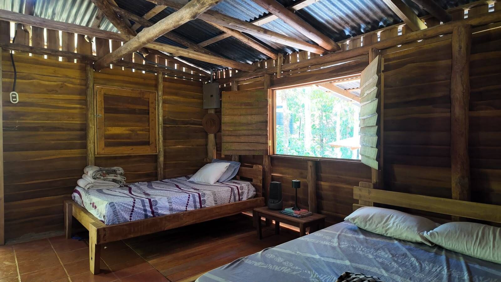 A wooden cabin with two beds, a small table, and a window with a view of trees.