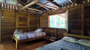 A wooden cabin with two beds, a small table, and a window with a view of trees.