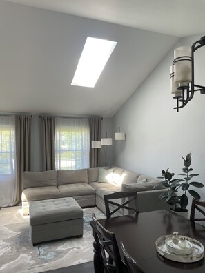 Ein modernes Wohnzimmer mit einem Ecksofa, einem Esstisch mit Stühlen, einer Pendelleuchte und einem Oberlicht.