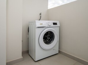 A white front-loading washing machine in a corner of a room with a tiled floor and a window. - Corporate Stays First Living - 102 (Asunción)