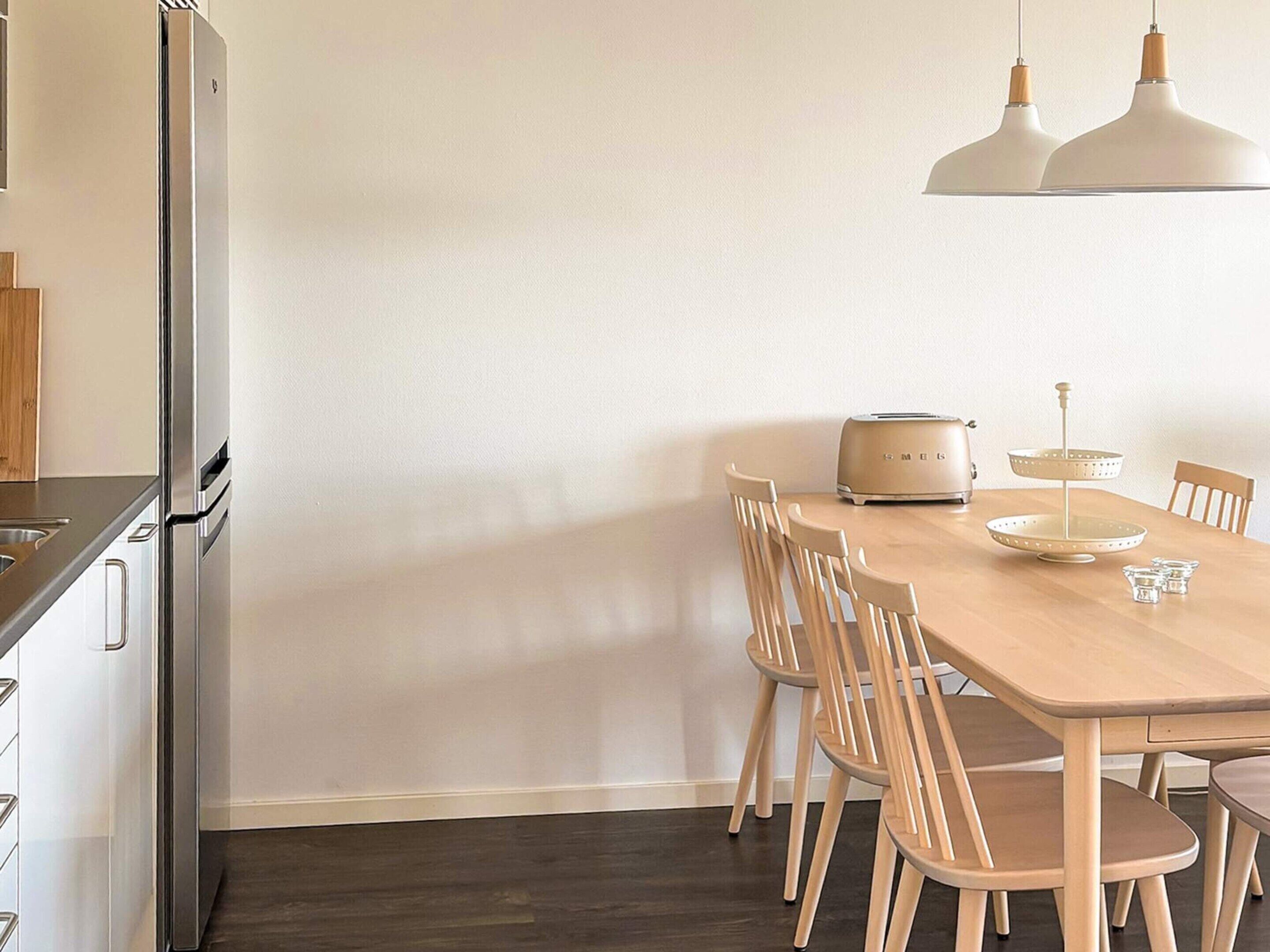 A modern kitchen with a stainless steel refrigerator, a wooden dining table with six chairs, and a two-light pendant ceiling.