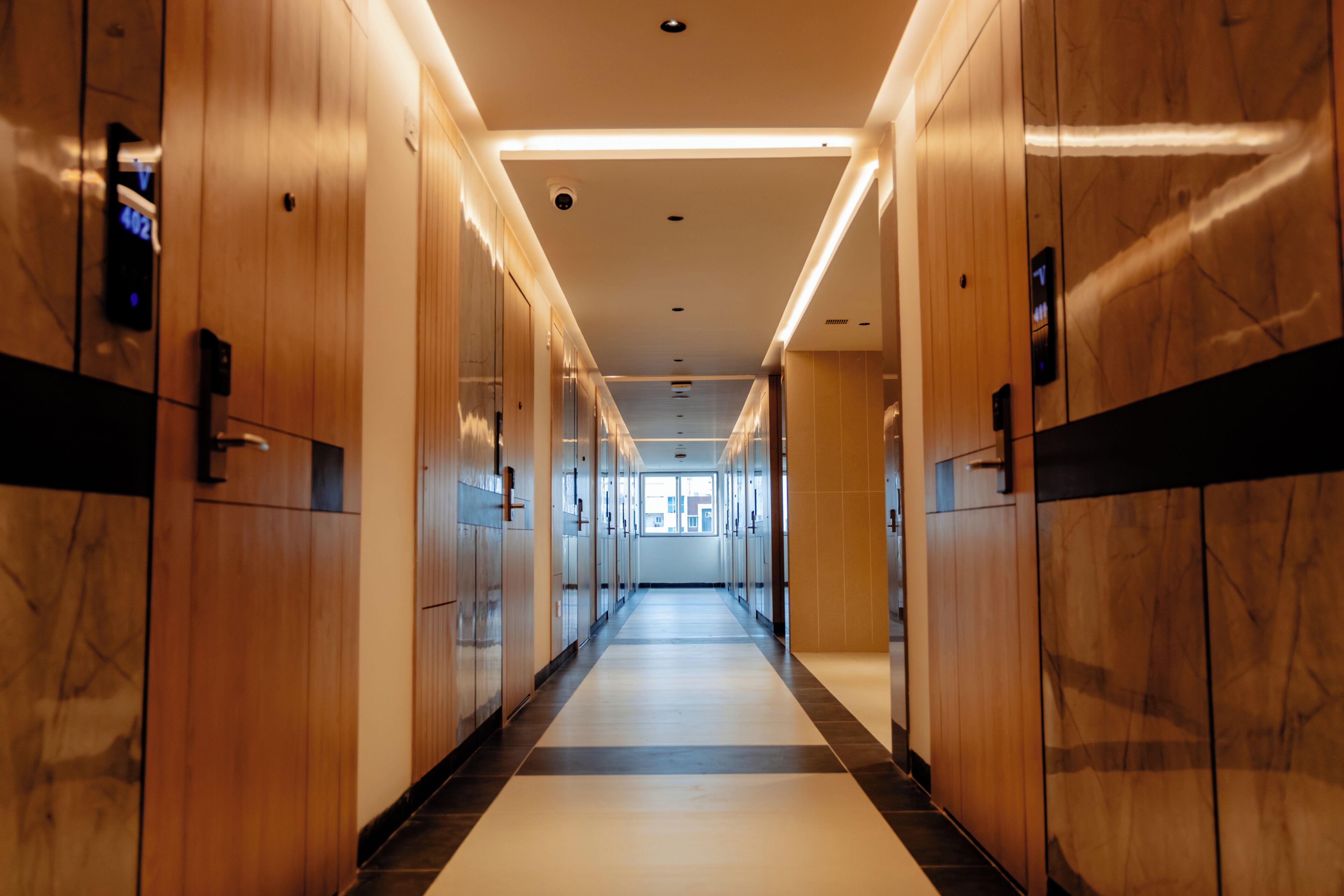 A long, narrow hallway with wooden paneling and a black and white tiled floor, leading to a bright area with windows.