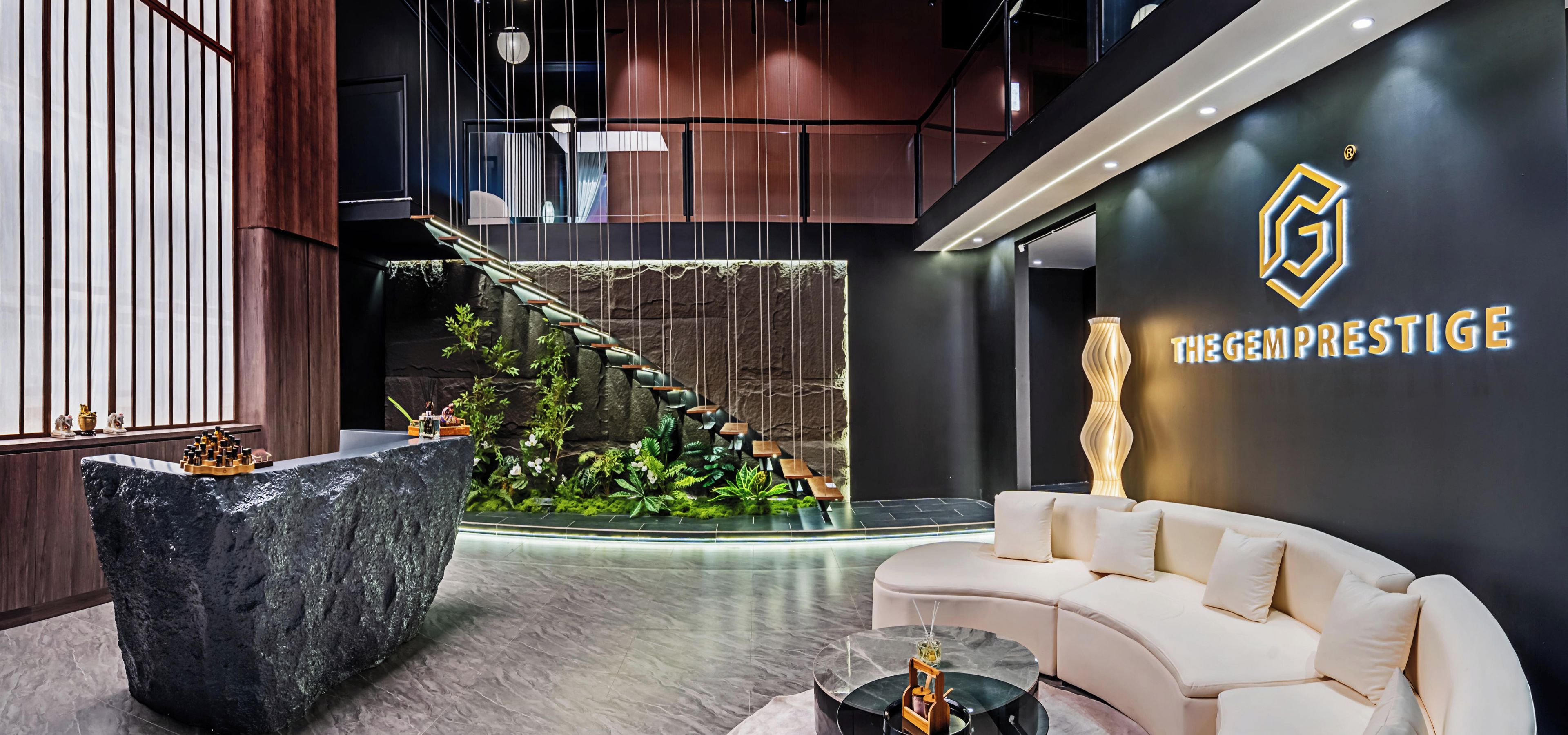 A modern lobby with a stone reception desk, a white sofa, a staircase with hanging chairs, and a wall with a logo and text.