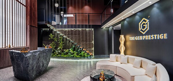 A modern lobby with a stone reception desk, a white sofa, a staircase with hanging chairs, and a wall with a logo and text.