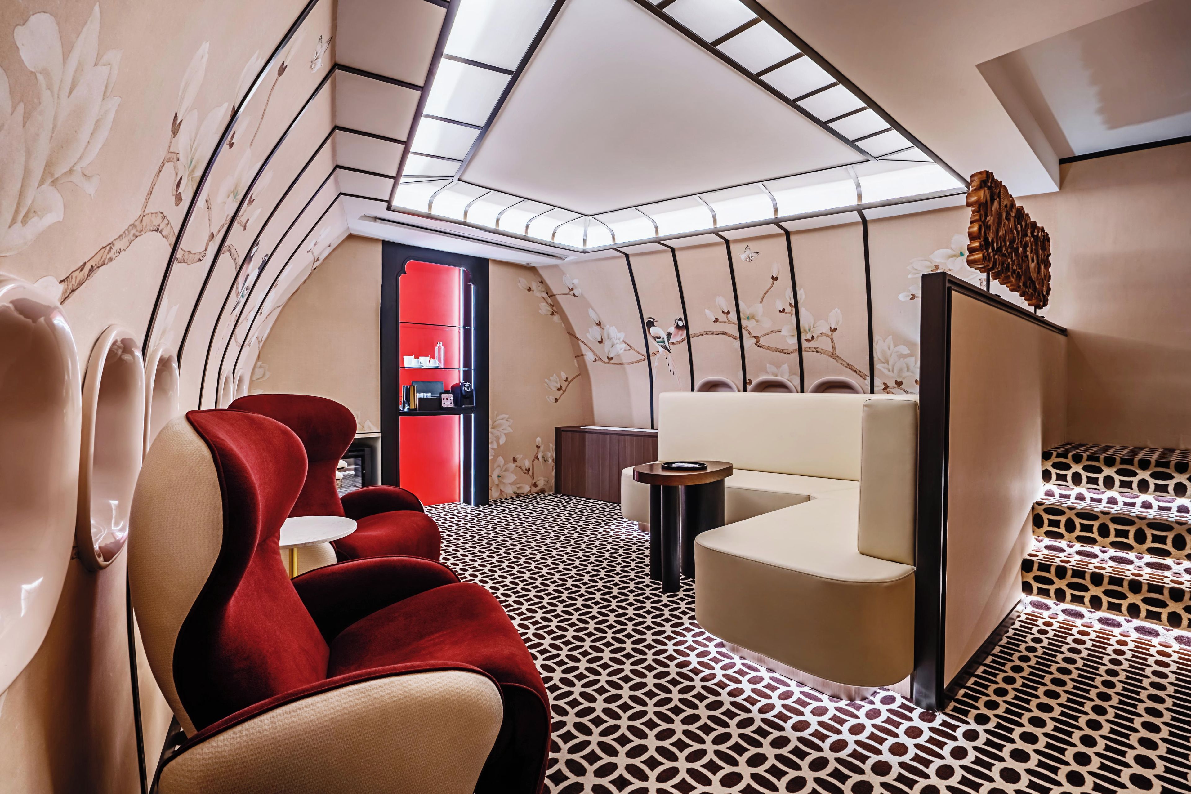 A modern interior with a sloped ceiling, red and beige seating, a patterned floor, and a red accent wall with a shelf.