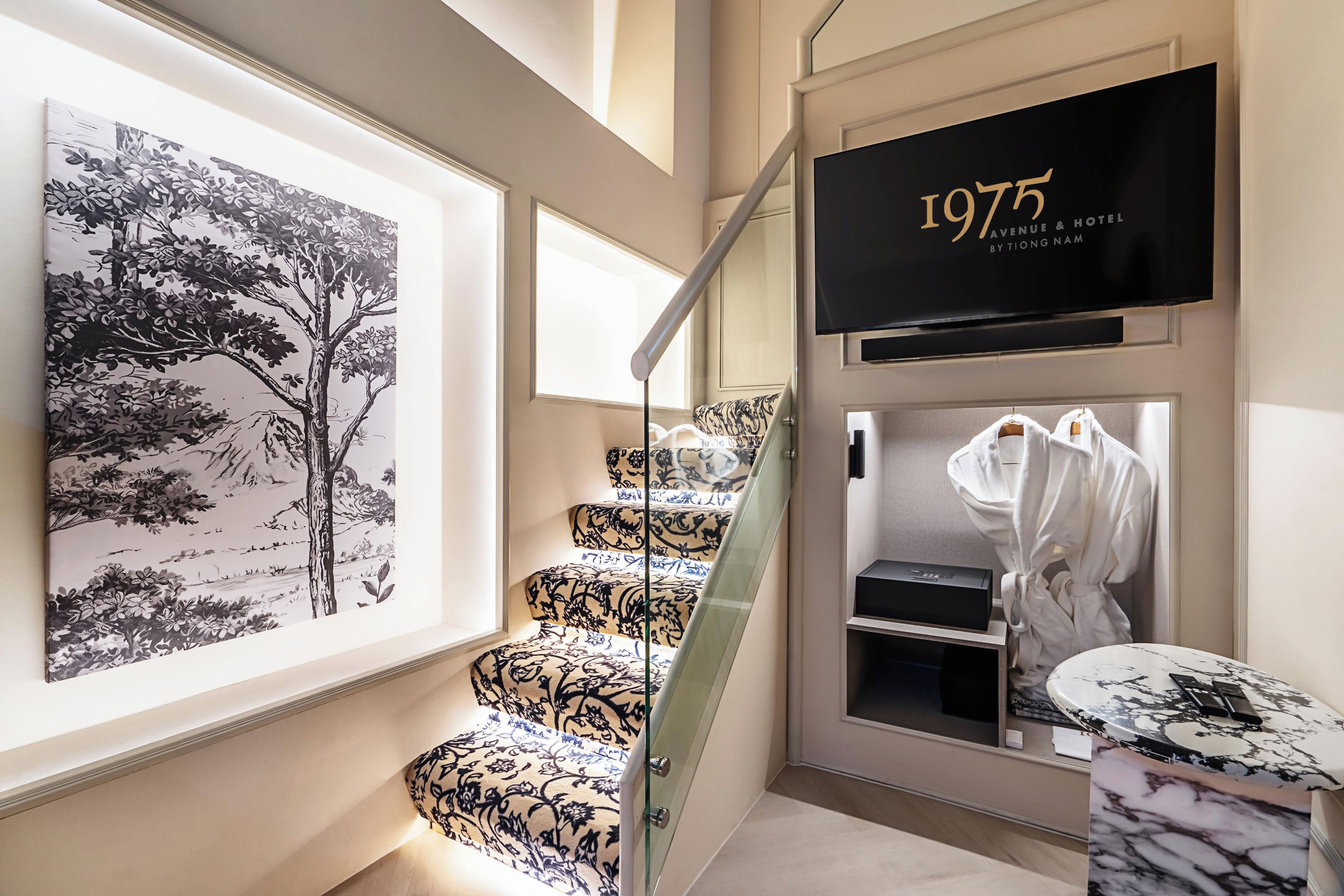 A staircase with patterned carpet, a framed black and white tree painting, a black sign with '1975 Avenue & Road by Tiong Nam', and a robe hanging on a hook.
