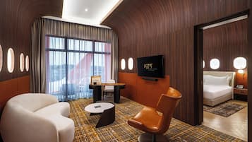 Ein modernes Hotelzimmer mit einer gewölbten Holzwand, einem großen Fenster mit Vorhängen, einem Flachbildfernseher, einem Sofa, einem Sessel und einem gemusterten Boden.