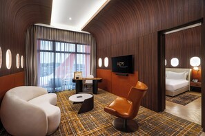 Une chambre d’hôtel moderne avec un mur en bois aux lignes courbes, une grande fenêtre avec des rideaux, un téléviseur à écran plat, un canapé, un fauteuil et un sol à motifs.