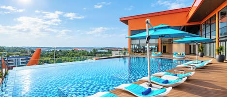 Área de piscina moderna de hotel com espreguiçadeiras e guarda-sóis, rodeada por um deck de madeira e vista para o horizonte urbano.
