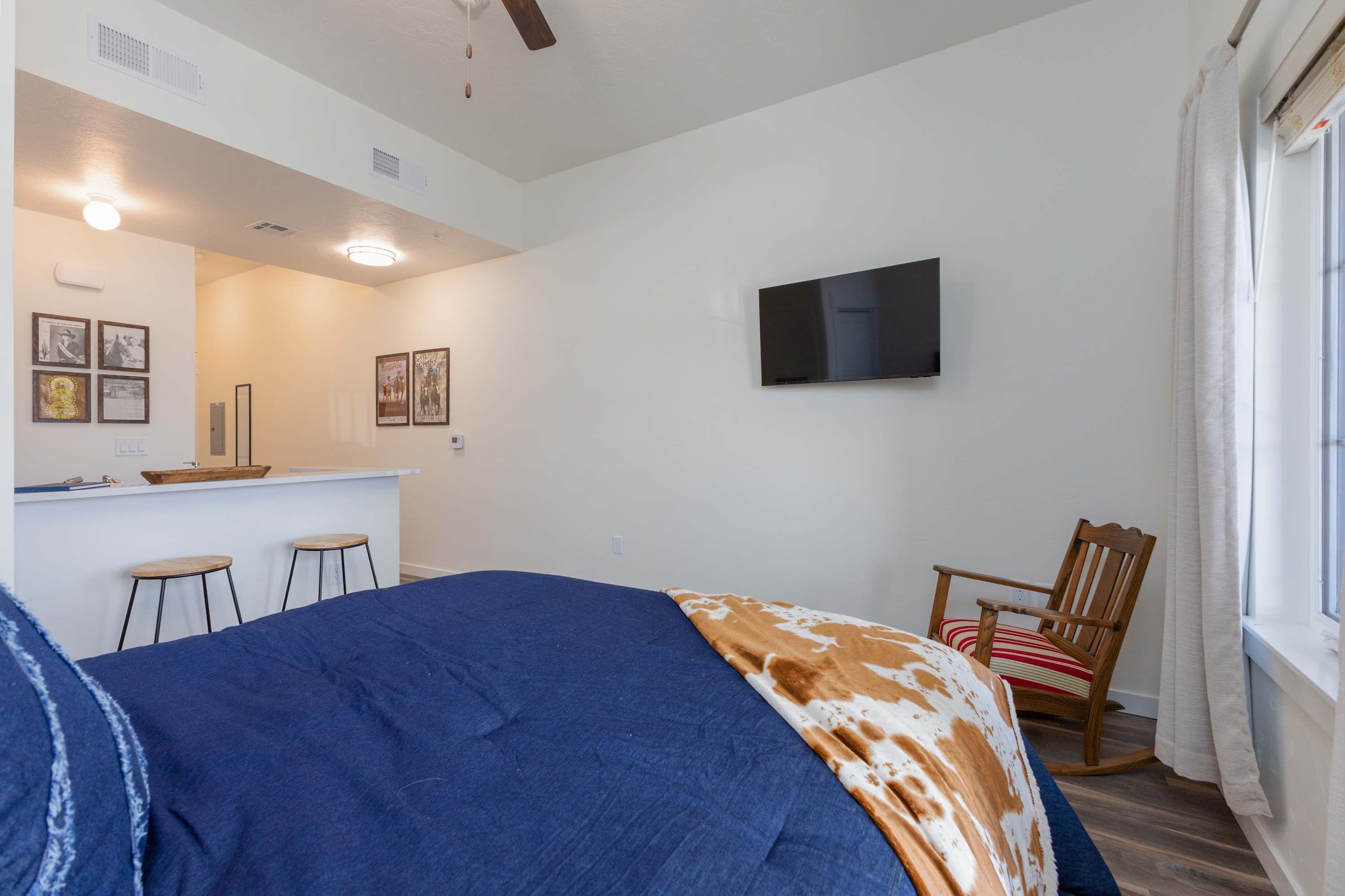 Un dormitorio con cama, una mecedora de madera, televisor montado en la pared y una zona de kitchenette.
