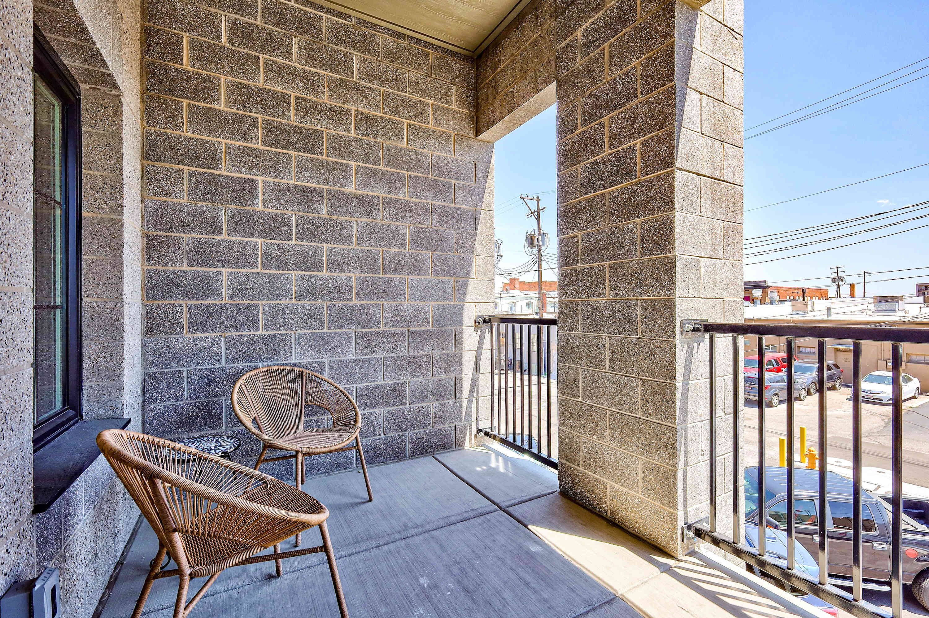 Un balcon avec un mur en briques, deux fauteuils en osier et une vue sur les voitures garées et les immeubles.