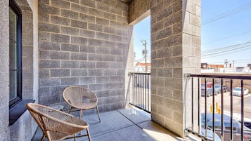 Un balcon avec un mur en briques, deux fauteuils en osier et une vue sur les voitures garées et les immeubles.