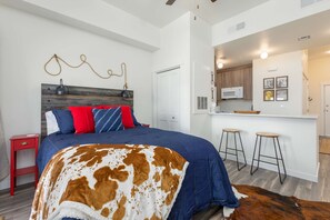 Ein Schlafzimmer mit einem Bett, einem roten Nachttisch, einer Kuhfelldecke, einem Holz-Kopfteil und einer Küchenzeile mit Hockern und einer Mikrowelle.