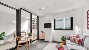 A modern living room with a sofa, a dining table with chairs, a TV mounted on the wall, and a large window. - Beach Petite - Across from Beach - Walk to City (Newcastle)