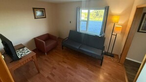 A living room with a sofa, a red chair, a wooden table, a lamp, and a window with a view. - Countryview Motel and Restaurant  (Steeves Mountain)