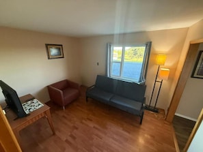 A living room with a sofa, a red chair, a wooden table, a lamp, and a window with a view. - Countryview Motel and Restaurant  (Steeves Mountain)