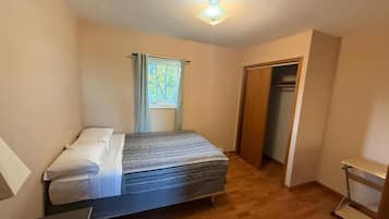 Un dormitorio con cama, ventana con cortinas, una puerta y una mesita.