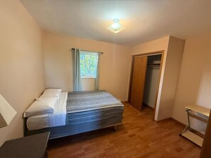 Un dormitorio con cama, ventana con cortinas, una puerta y una mesita.