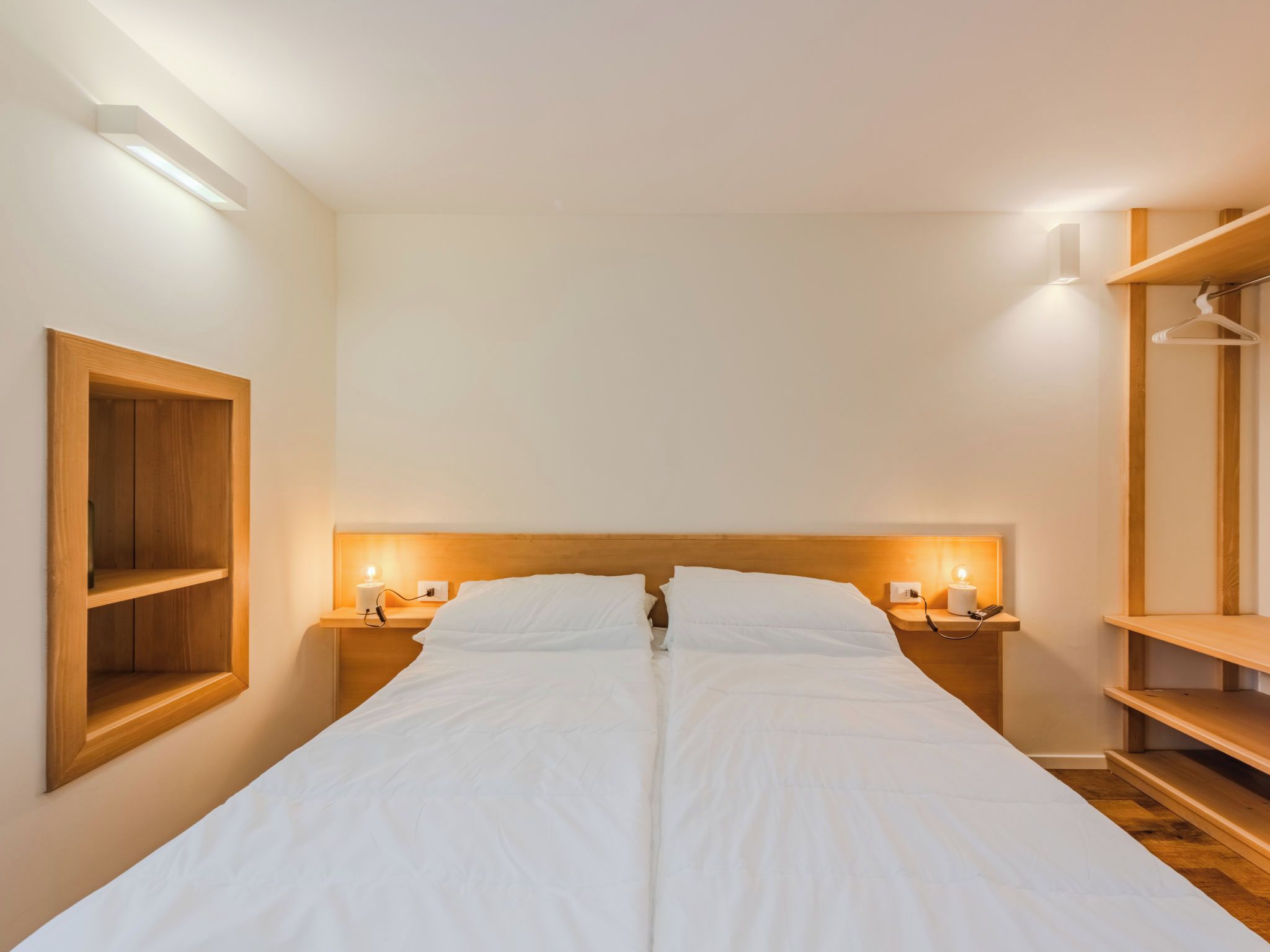 A bedroom with a wooden headboard, two bedside tables with lamps, a wooden shelf unit, and a wooden floor.