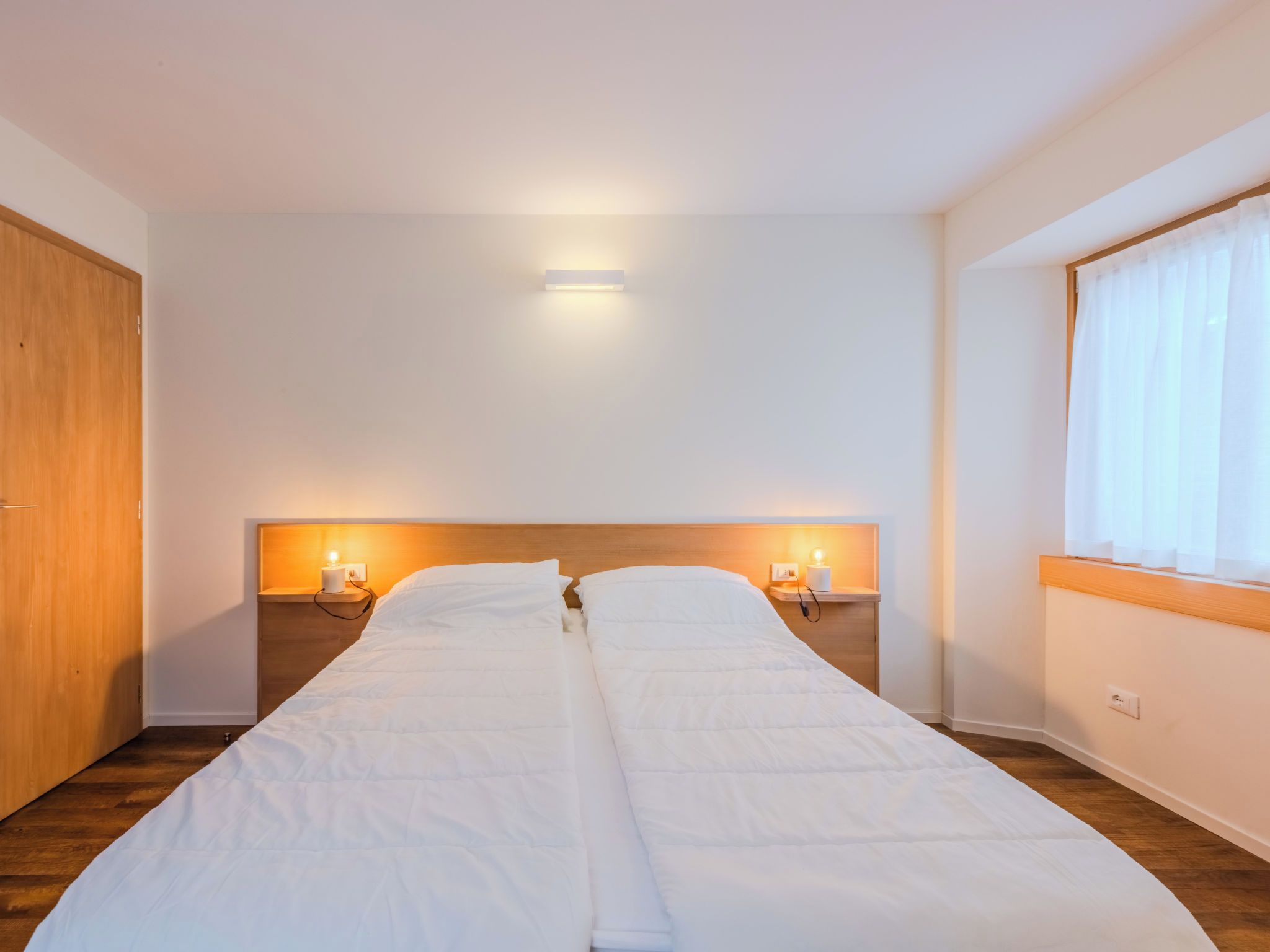 Cama doble con ropa de cama blanca, dos lámparas de noche, cabecera de madera y una ventana con cortinas de encaje.
