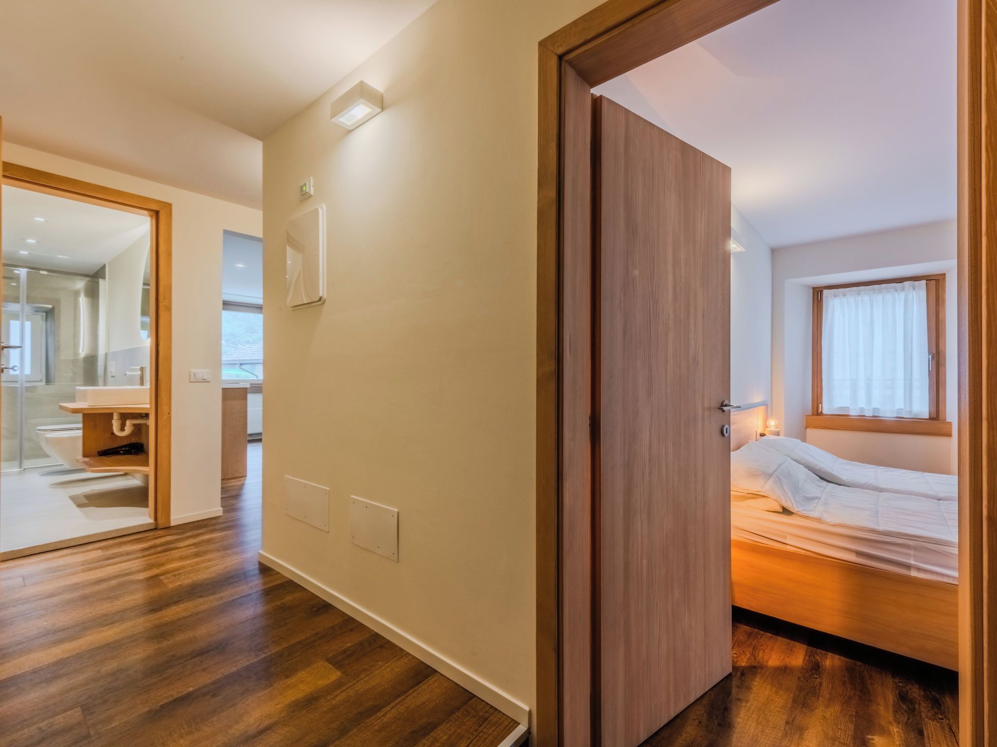 Ein Hotelzimmer mit Holzboden, einem Bett mit weißer Bettwäsche, einem Fenster mit Vorhängen und einem durch eine offene Tür einsehbarer Badezimmer.