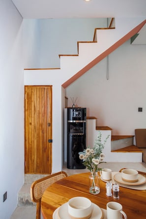 Uma sala de jantar com uma mesa de madeira posta para quatro pessoas, um vaso com flores e um eletrodoméstico preto ao fundo.
