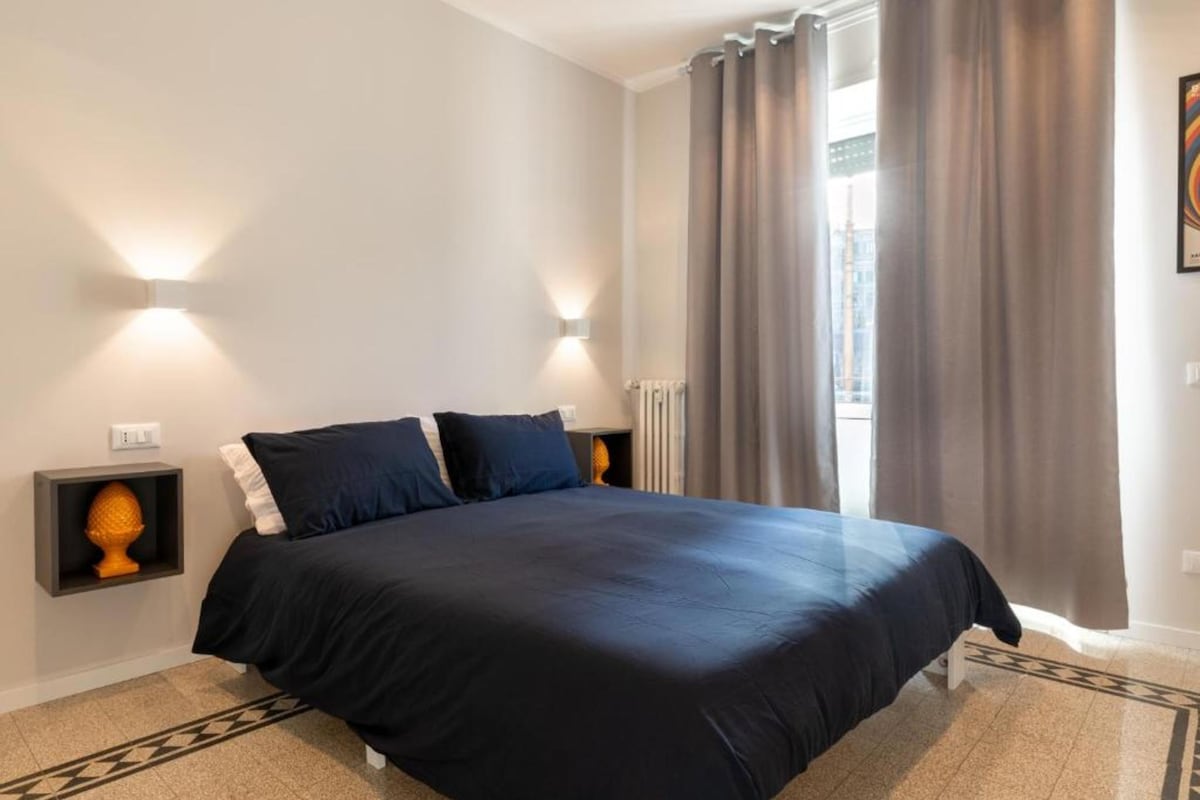 A bedroom with a bed, two bedside tables, a radiator, and a window with curtains.