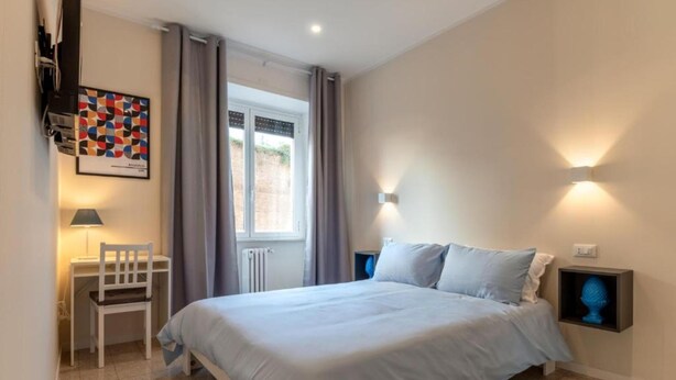 A bedroom with a bed, a chair, a desk, a window, and a wall-mounted light fixture. - Brick & Arch (Roma)