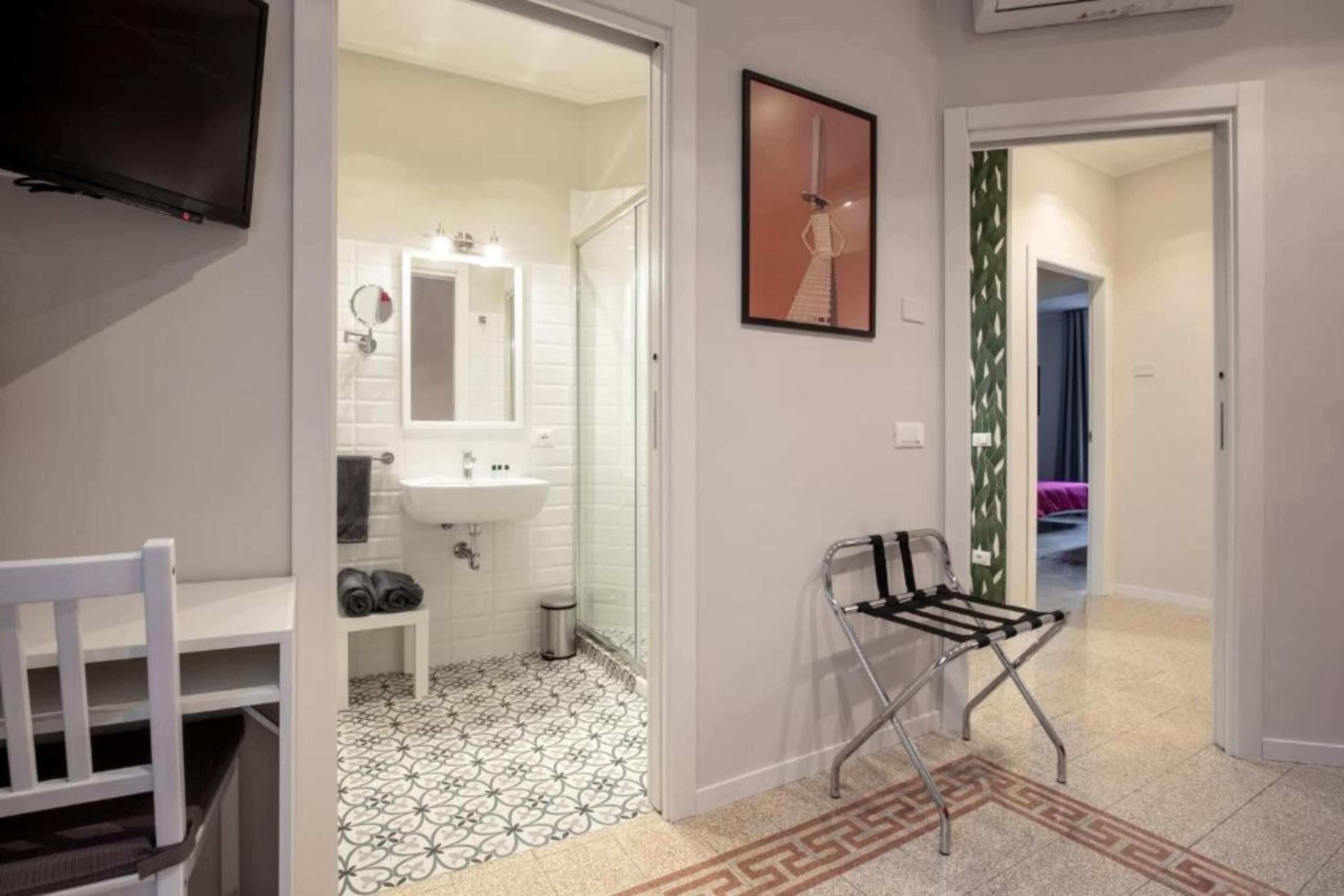 A hotel room with a white chair, a small table, a bathroom with a sink and mirror, and a patterned floor.