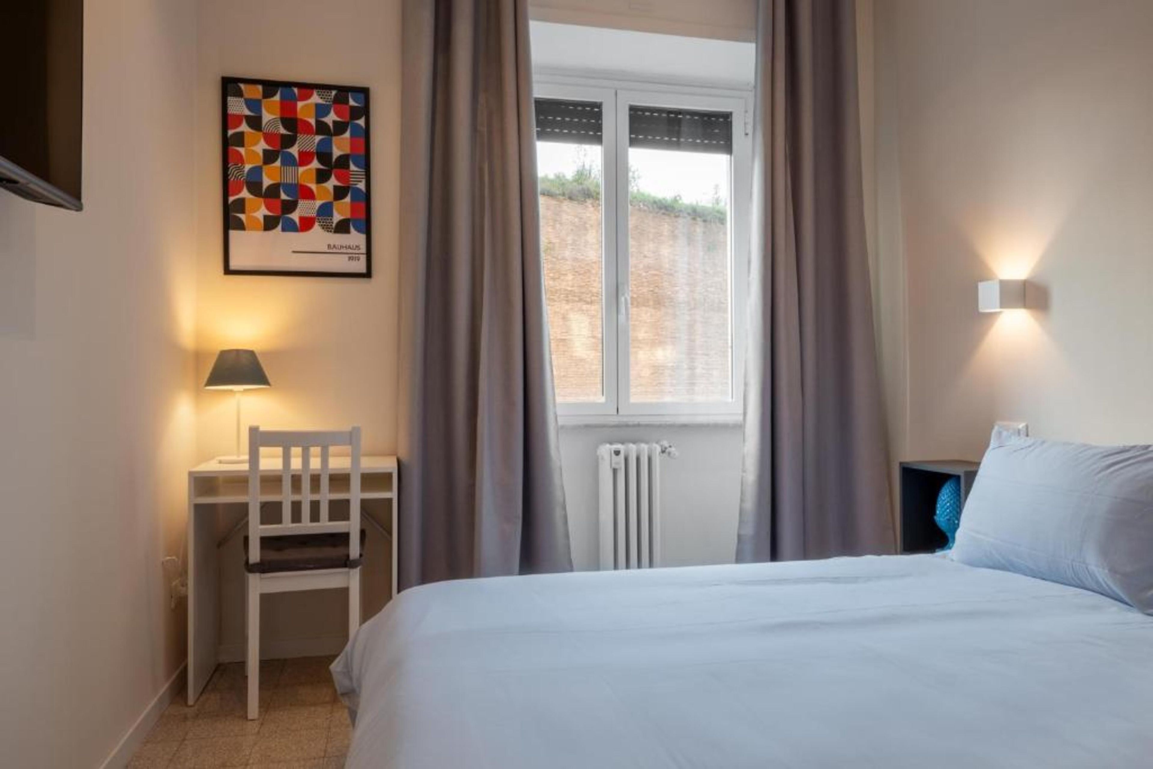 Ein Hotelzimmer mit Bett, Schreibtisch mit Stuhl, Fenster mit Vorhängen und einer Wandleuchte.