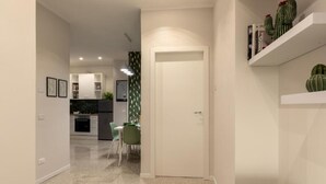 A modern kitchen with white cabinets, a dining area with a green chair, and a white door leading to another room. - Brick & Arch (Roma)