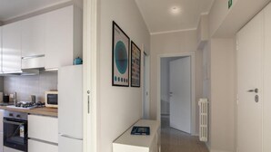 A modern kitchen with white cabinetry, a microwave, and an oven. There is a hallway with a radiator and a door leading to another room. - Brick & Arch (Roma)
