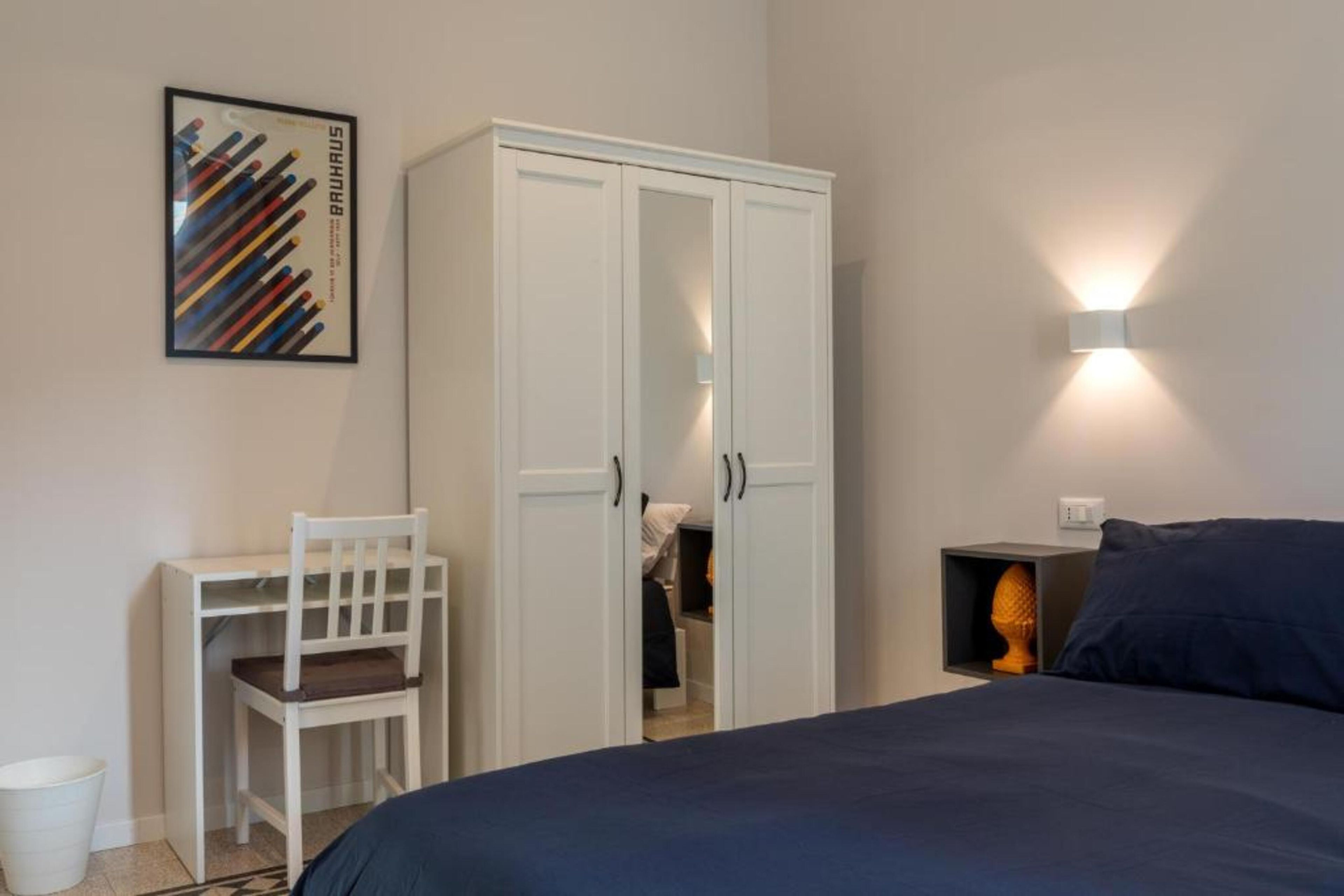 A bedroom with a bed, a wardrobe, a desk with a chair, and a wall-mounted light fixture.