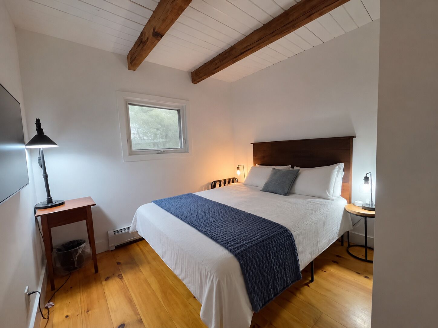 Quarto com cama, mesa de cabeceira, luminária, janela e vigas de madeira no teto.
