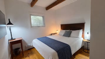 Quarto com cama, mesa de cabeceira, luminária, janela e vigas de madeira no teto.