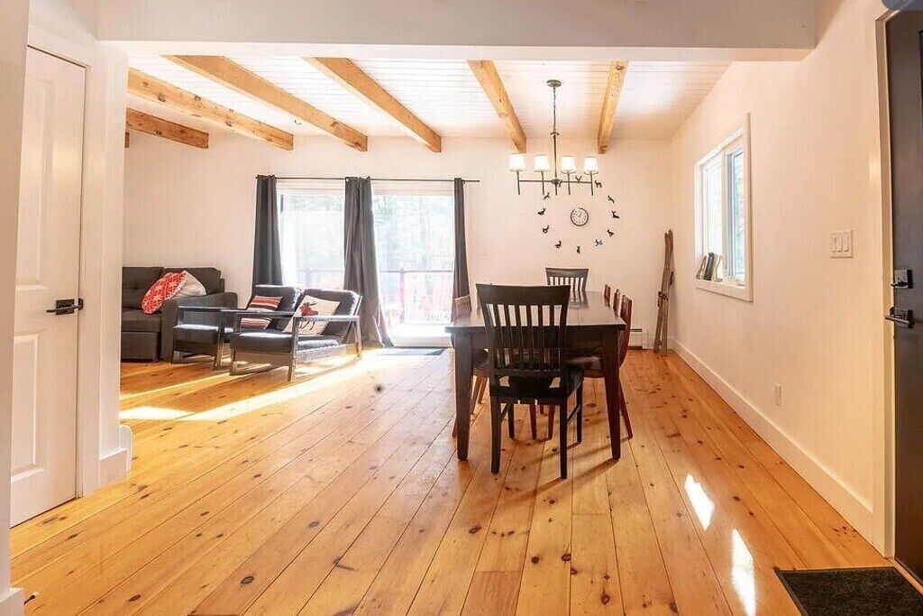 Ein Zimmer mit Holzboden, einem Esstisch mit Stühlen, einem Sofa, einer Kristallleuchtre und einem fenster mit Vorhängen.